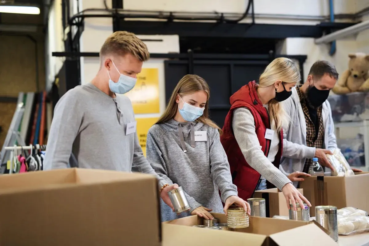 Four volunteers in face masks pack canned food into cardboard boxes.